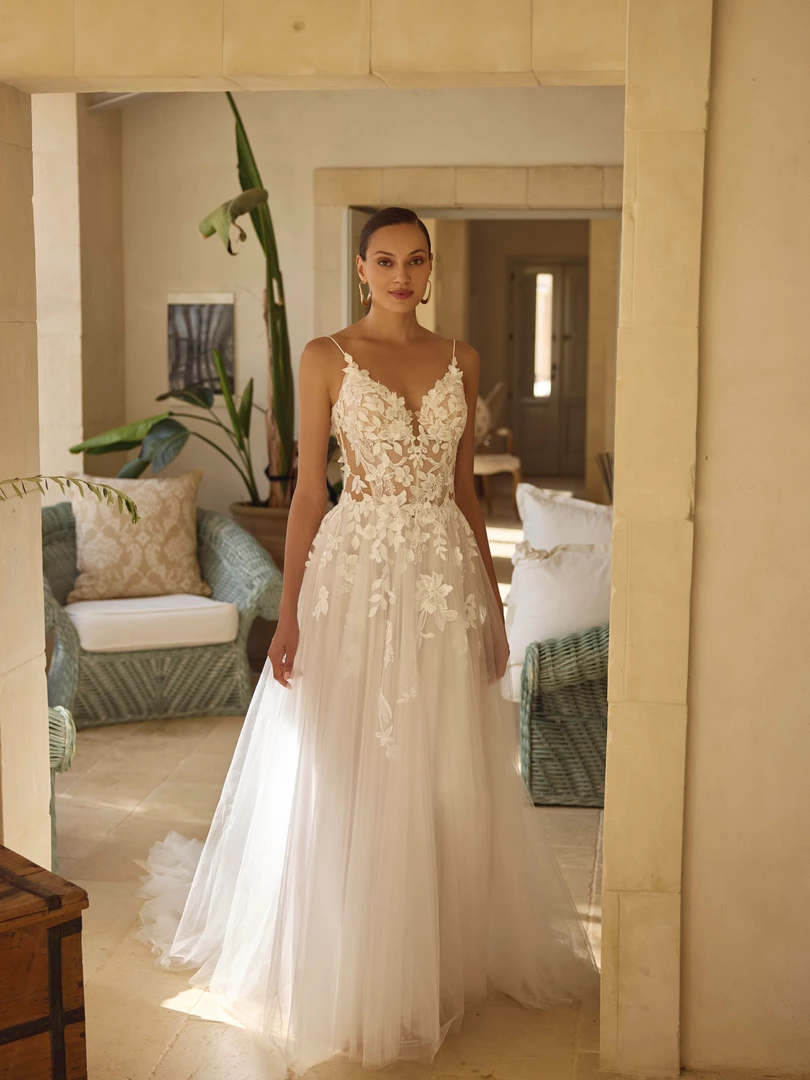 Braut im ärmellosen Kleid mit tiefem V-Ausschnitt und floralen Spitzenapplikationen in einem Türrahmen.